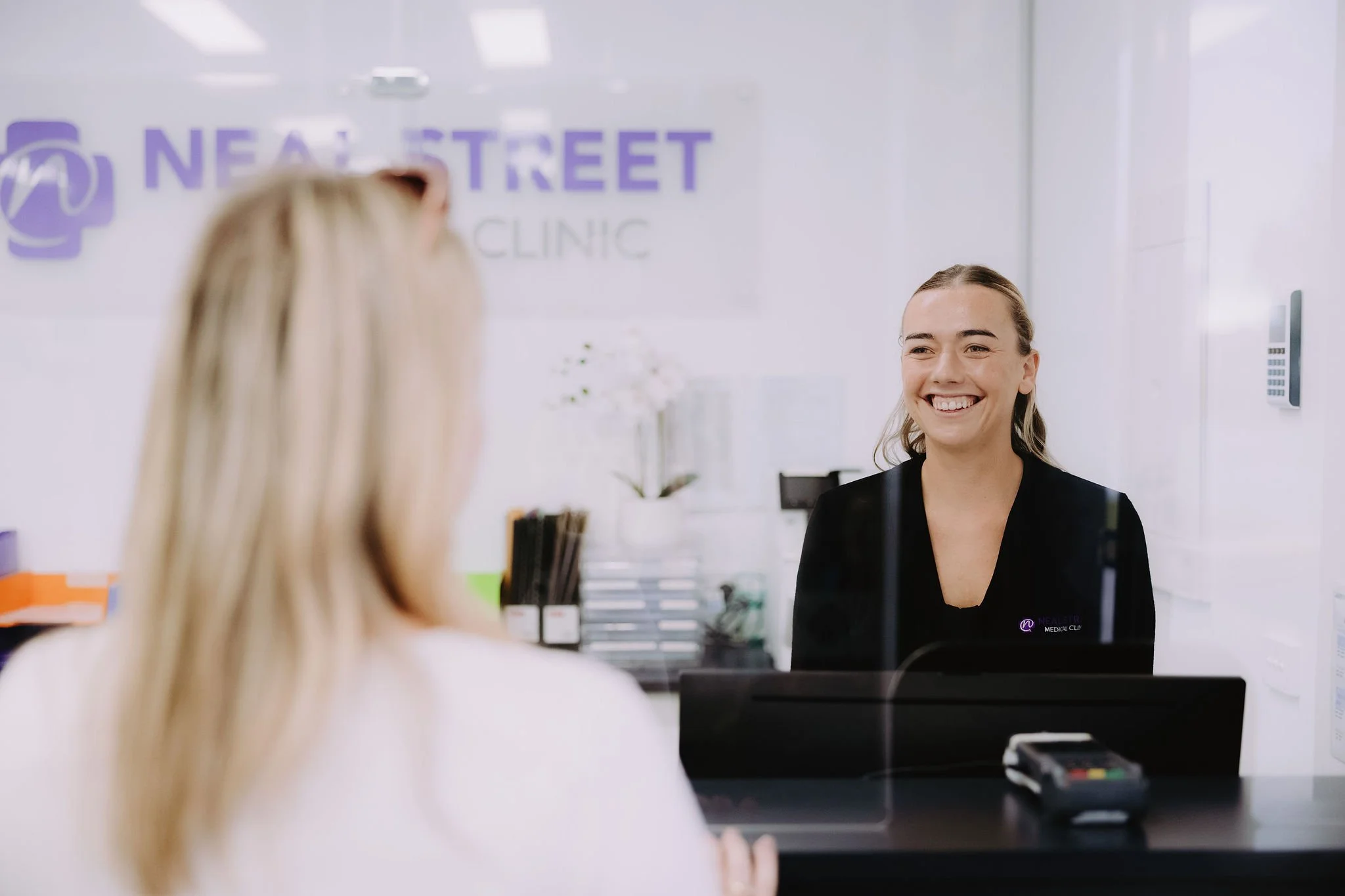 Reception area at a medical clinic with a smiling receptionist behind the counter, visible sign on the wall reading 'NEAL STREET CLINIC'.