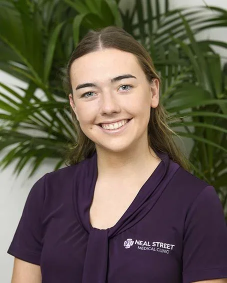 A young woman with brown hair and a purple shirt, smiling, standing in front of green tropical plants.