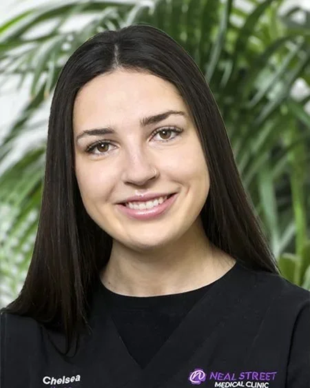 A young woman with long dark hair, smiling, wearing a black shirt with a medical clinic logo, in front of green leafy plants.