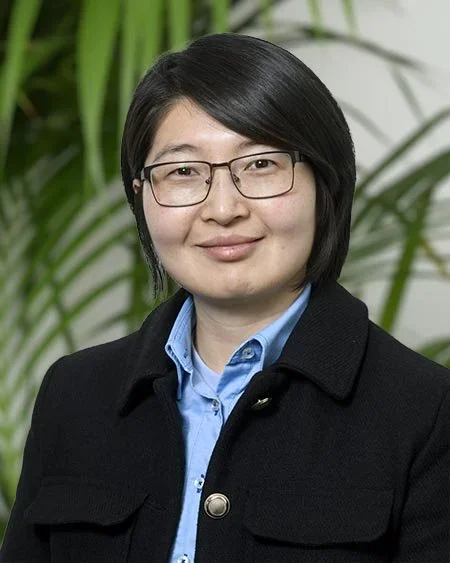 A woman with short black hair and glasses, wearing a blue shirt and black jacket, standing outdoors with green plants in the background.