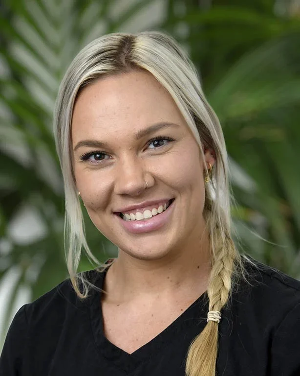 Smiling woman with blonde braided hair, wearing a black top, in front of green foliage.