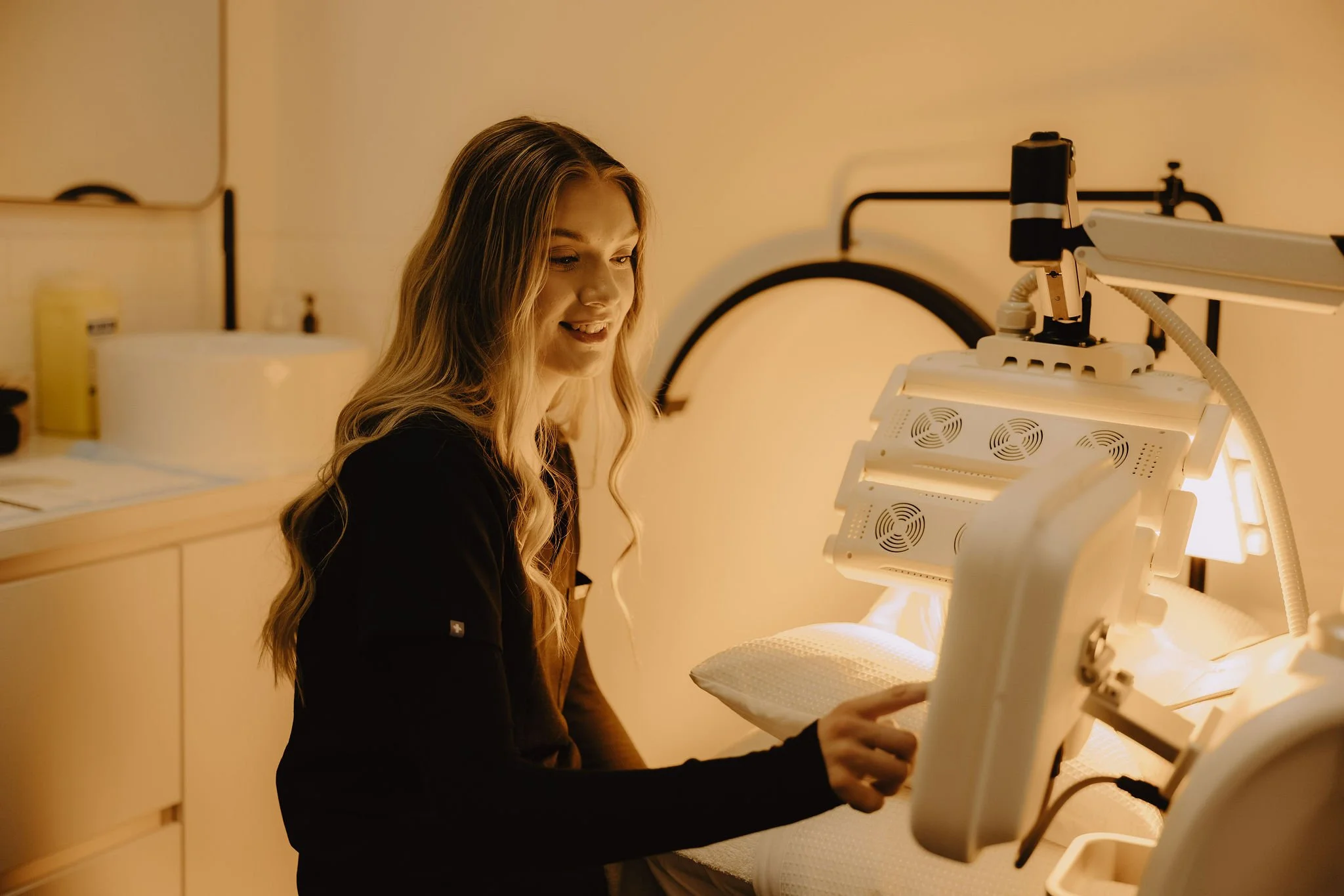 A woman with long blonde hair smiling while lying in a medical bed, touching a medical device with her finger, in a warmly lit room.
