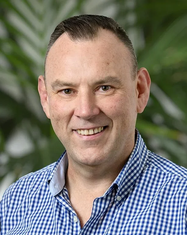 A smiling man with short, dark hair wearing a blue and white checkered shirt, standing in front of a background with green foliage.