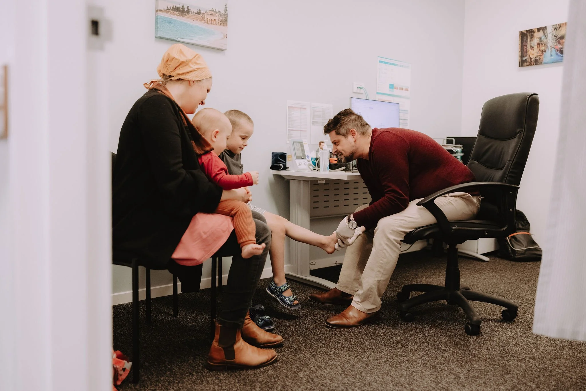 A doctor examining a child's foot while three children and a woman watch inside a medical office.