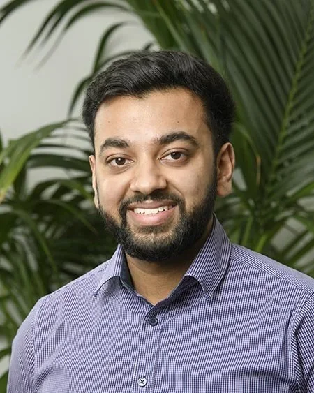 A man with dark hair and a beard smiling in front of green plants, wearing a purple checkered button-up shirt.