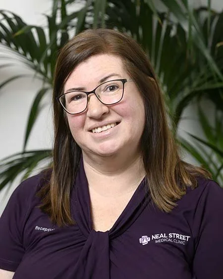 A woman with brown hair and glasses smiling in front of green plants, wearing a black uniform with a Neal Street Medical Clinic logo.