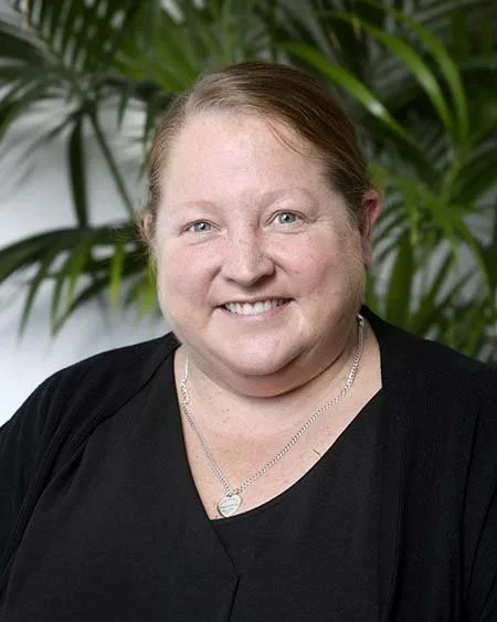 Portrait of a smiling woman with light skin, brown hair, and blue eyes, wearing a black top and a silver necklace, standing in front of green palm leaves.