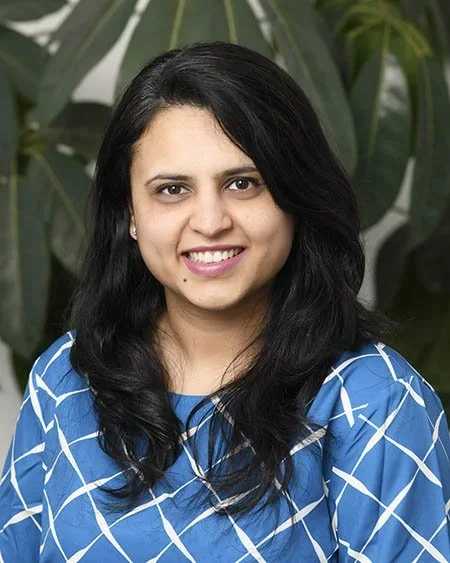 A woman with long black hair wearing a blue patterned blouse standing in front of green plants.