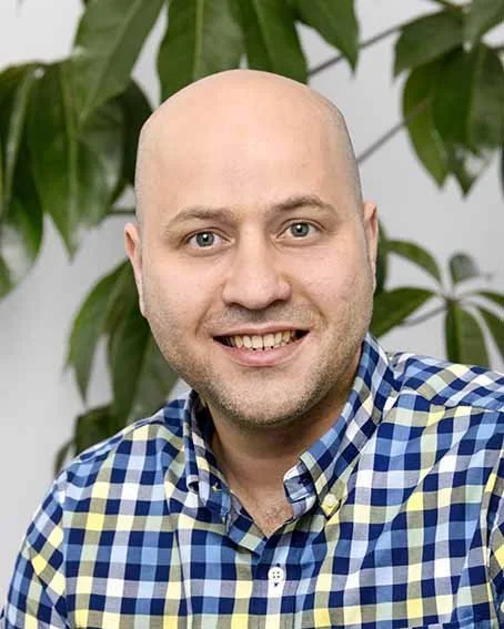 Portrait of a smiling man with a shaved head, wearing a blue and yellow checkered shirt, standing in front of green plants.
