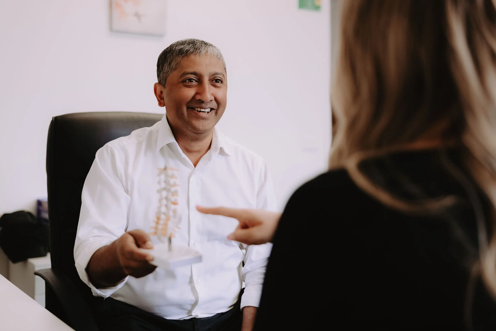 A man in a white shirt holding a small model of a spinal column, smiling while talking to a woman in a black top in an office setting.