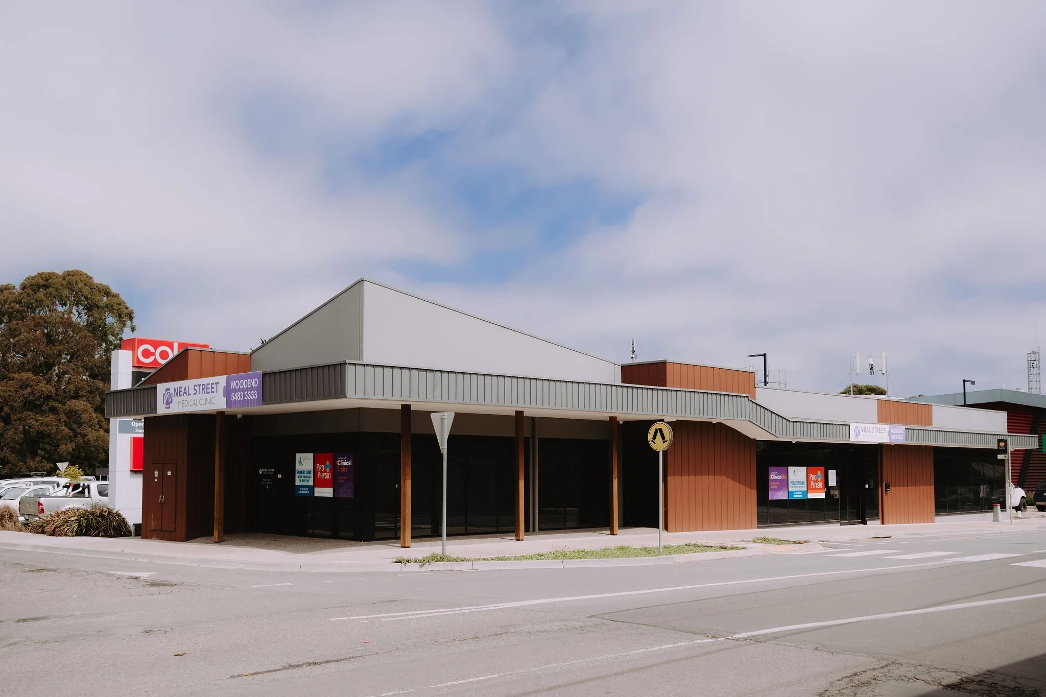 Modern commercial building with signage for Neal Street Medical Clinic and Woodend. Located on a street corner with a parking lot and a traffic sign visible.