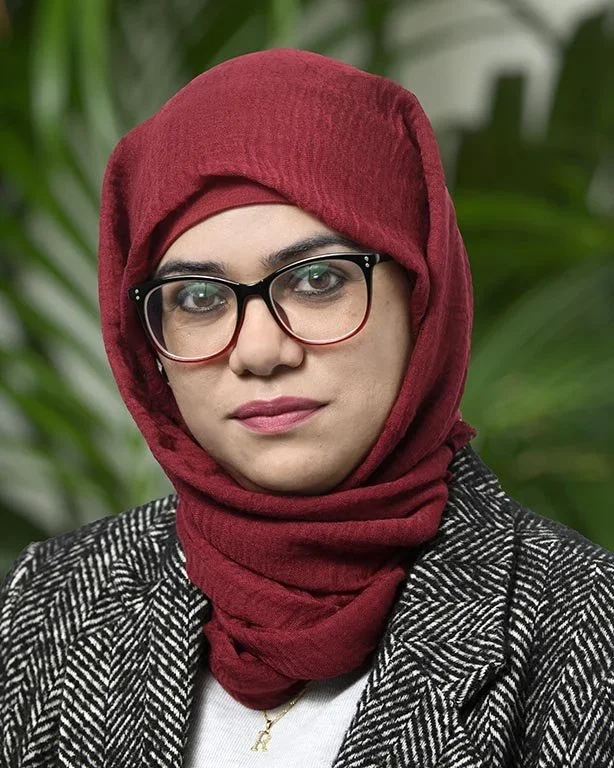 A woman wearing a red headscarf, black glasses, and a patterned blazer, standing outdoors with green foliage in the background.