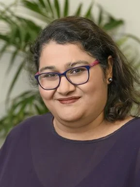 A woman with dark hair and glasses smiling in front of a green leafy plant.