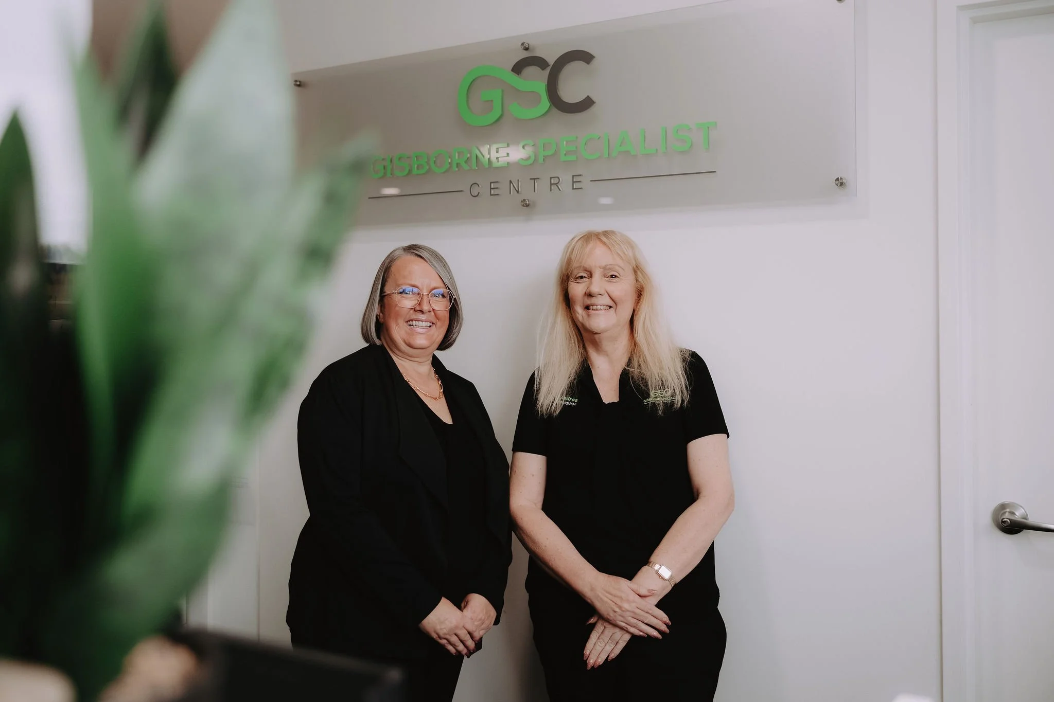 Two women standing in front of a sign that reads "GSC Gisborne Specialist Centre" at an indoor healthcare facility. One woman has short gray hair with glasses, the other has long blonde hair. Both are smiling and wearing black clothing.