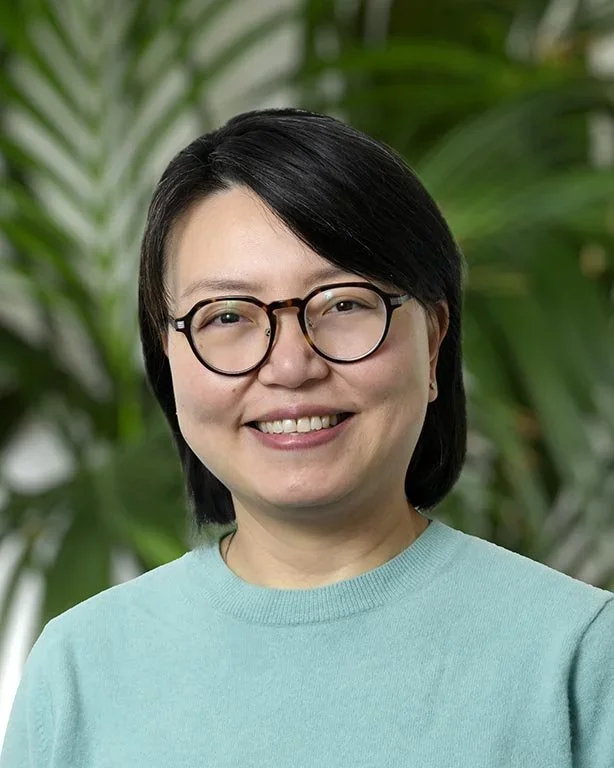 Smiling woman with short black hair, wearing glasses and a light blue sweater, standing in front of green plants.