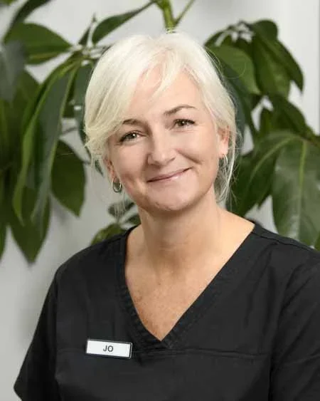 A smiling woman with short white hair, wearing a black uniform with a name tag that says 'Jo', standing in front of green leafy plants.
