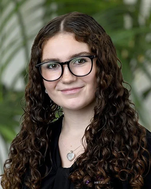 A young woman with curly brown hair, wearing black glasses and a black top, stands in front of a green leafy background.