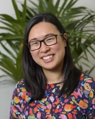 A woman with black hair, glasses, and a colorful patterned top, smiling, with large green plants in the background.