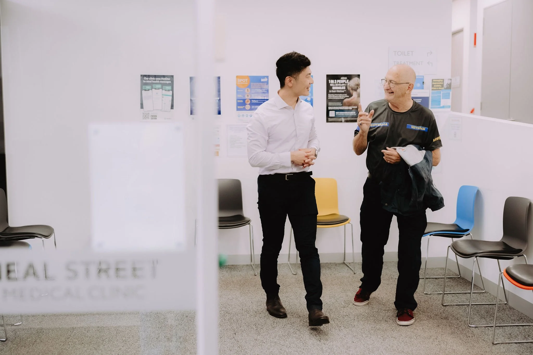 Two men are talking and smiling in a waiting room with chairs. One is a young man in a white shirt and black pants, and the other is an older man in a black t-shirt with patches, holding a jacket and paper.