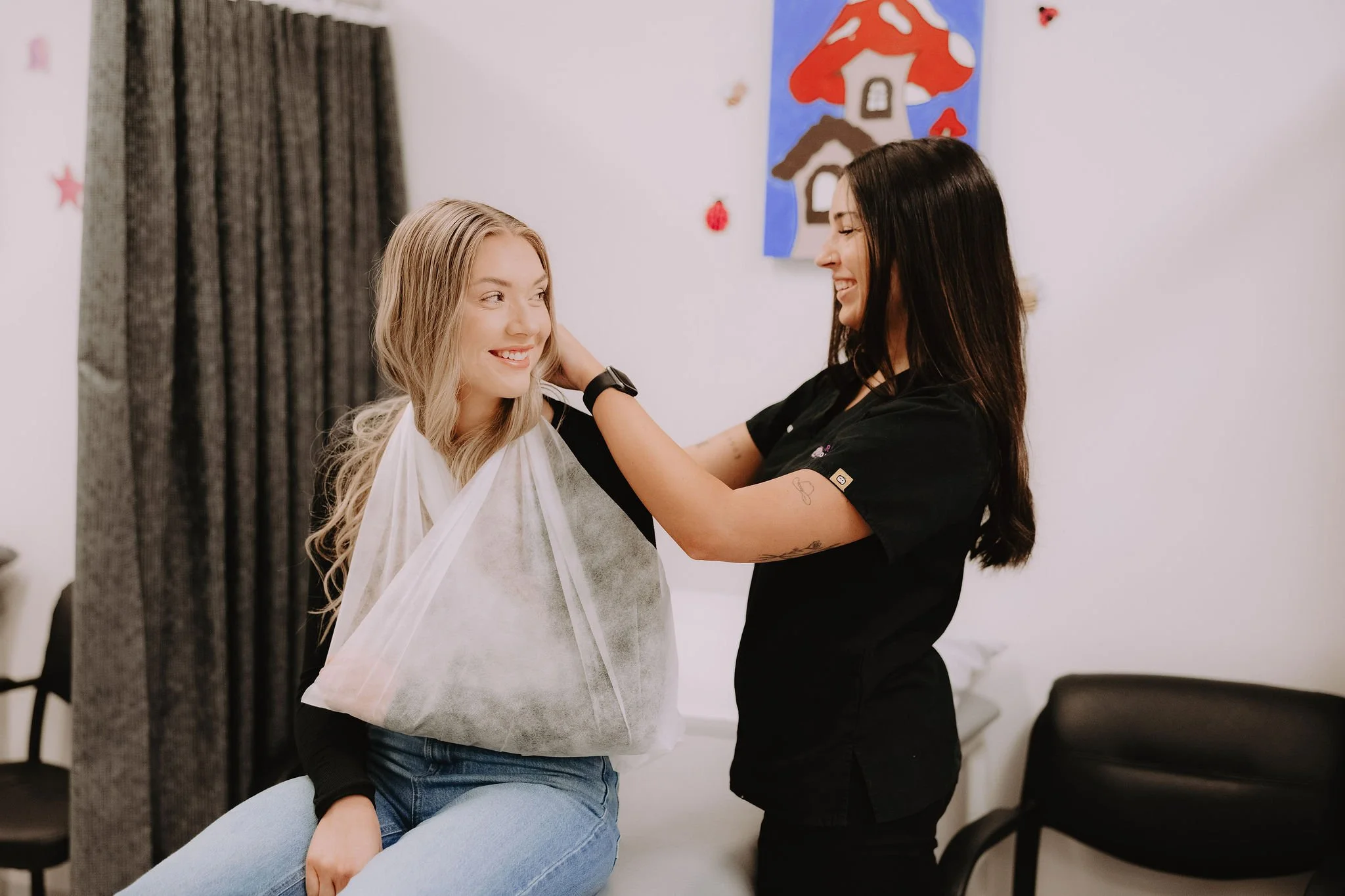 A woman with blonde hair sits on a chair, smiling, as a hairstylist with long dark hair styles her hair. They are in a room with light-colored walls decorated with colorful artwork and small decorations.