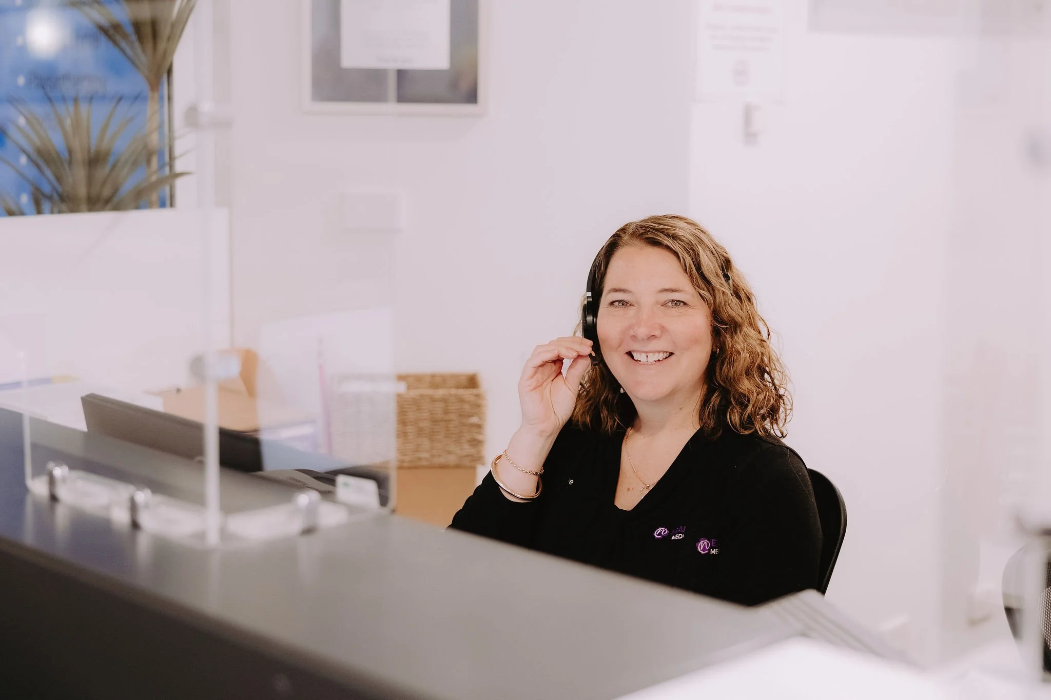 A smiling woman with curly hair, wearing a black top and headphones, sitting at a reception desk in an office environment.