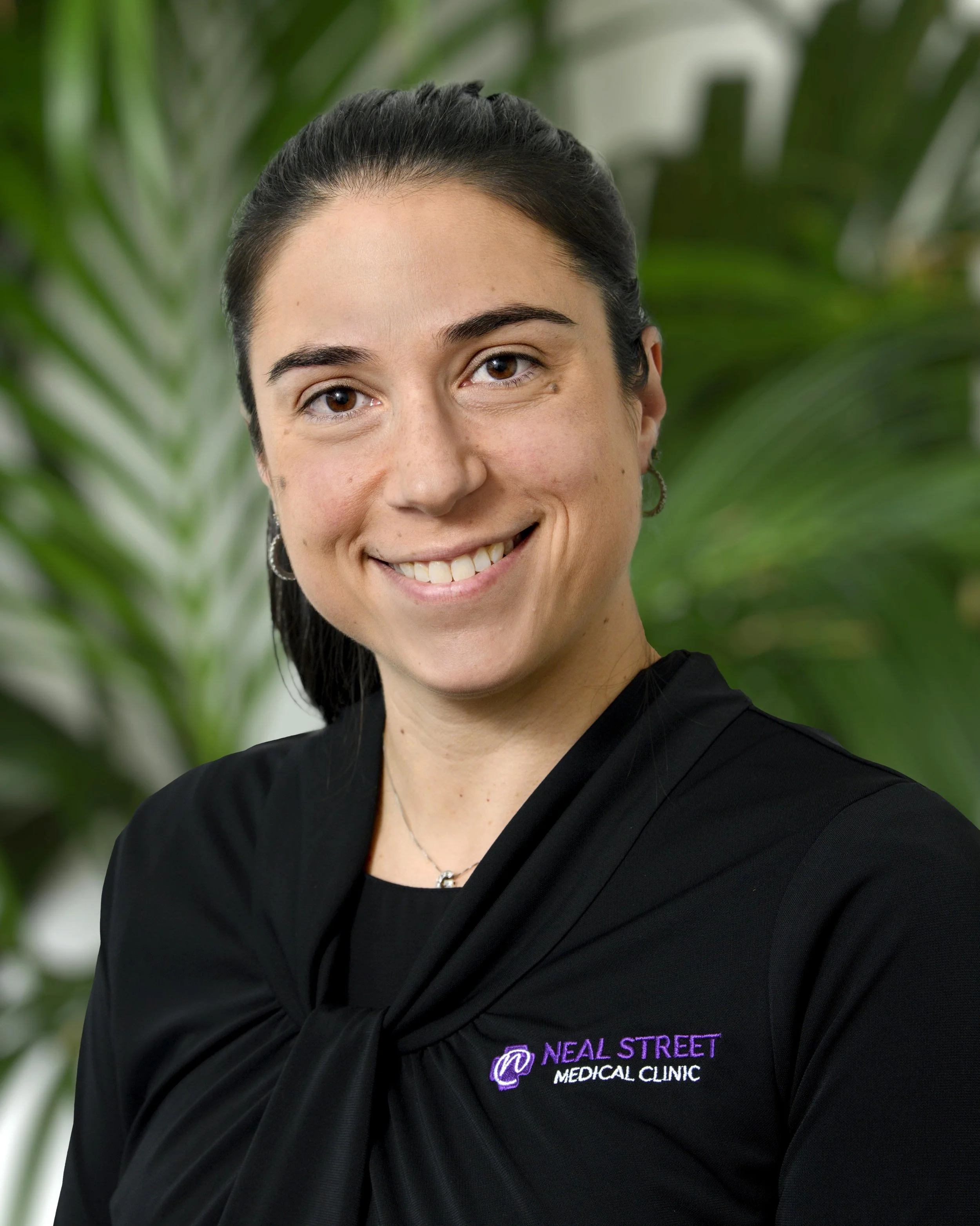A young woman with brown hair and a purple shirt, smiling, standing in front of green tropical plants.