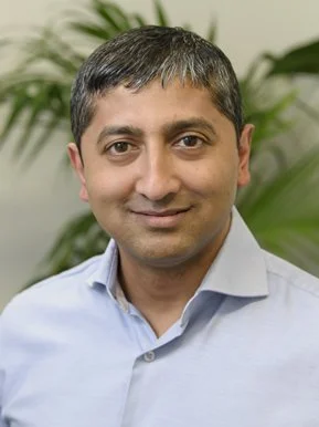 Headshot of a man with short dark hair, wearing a light blue collared shirt, standing in front of green plants.