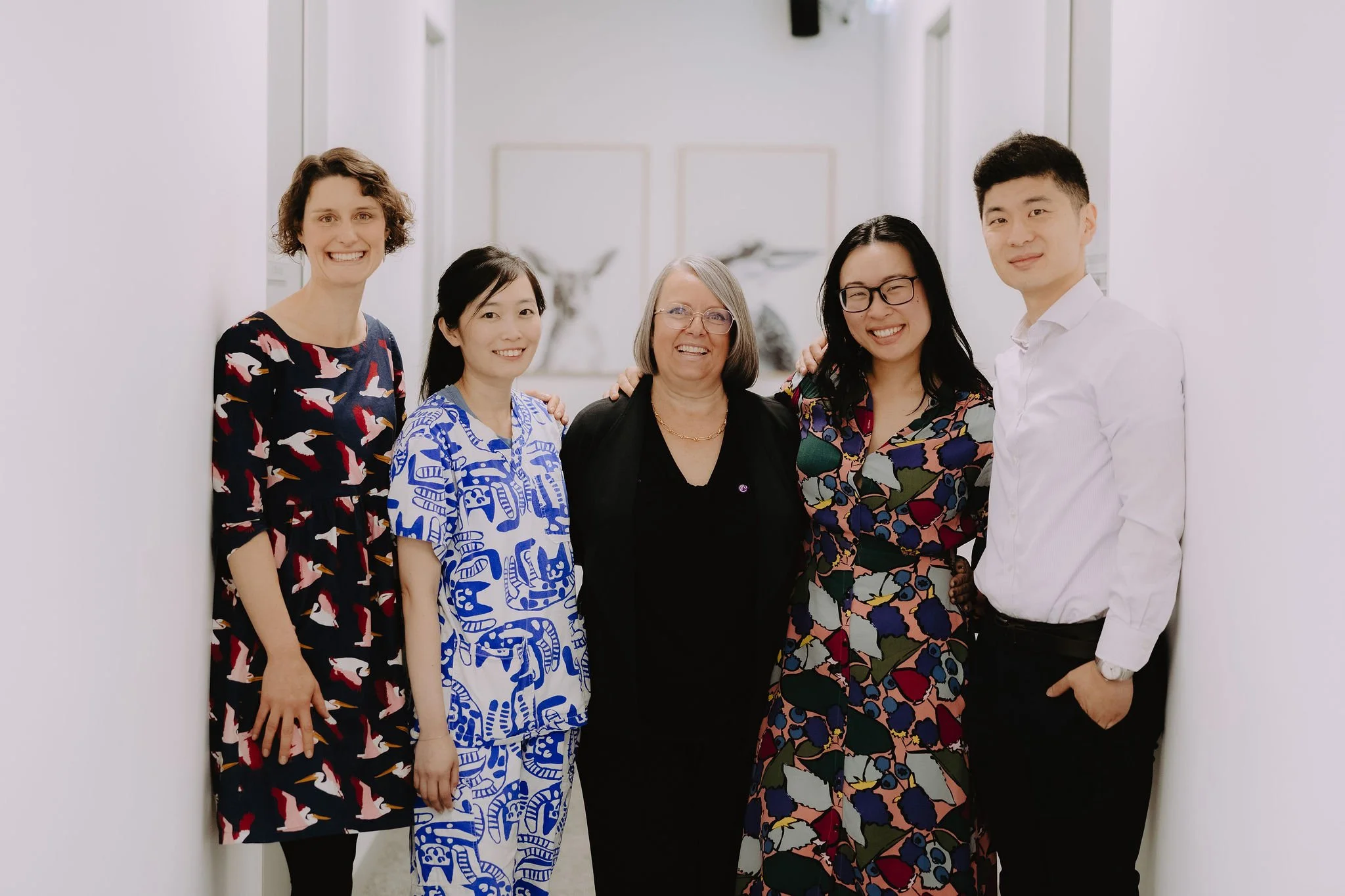 Group of five diverse women standing together in a bright hallway, smiling at the camera, dressed in colorful and patterned outfits.