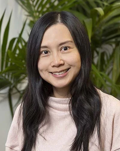 A woman with long dark hair smiling in front of a background of green plants.