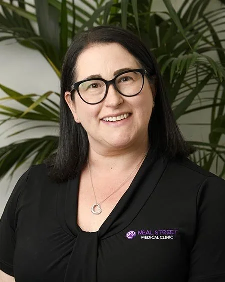 A woman with dark hair, glasses, and a black top with a logo that says 'Neal Street Medical Clinic,' standing in front of green plants.