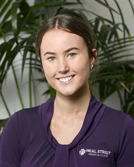 A young woman with brown hair wearing a purple shirt with a Neal Street Medical Clinic logo, standing in front of green plants.
