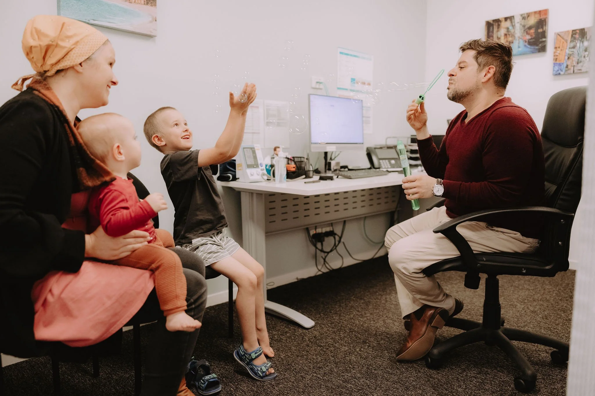 A doctor or therapist in an office with three children. The doctor is sitting in a black office chair, wearing a maroon sweater and beige pants, blowing bubbles as the children watch and laugh.