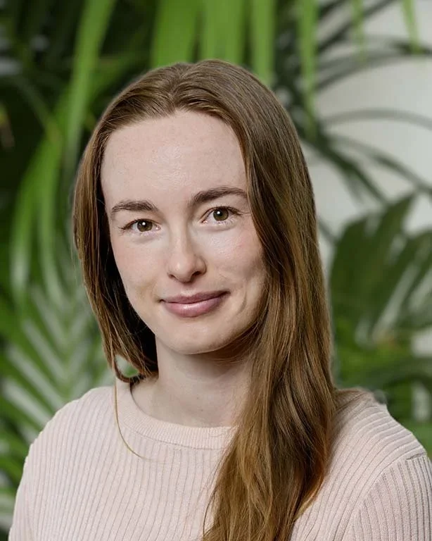 A woman with long red hair and fair skin smiling at the camera, wearing a light pink sweater, with large green tropical leaves in the background.