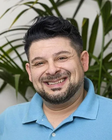 A smiling man with dark hair and a beard, wearing a blue polo shirt, is standing in front of green plants.