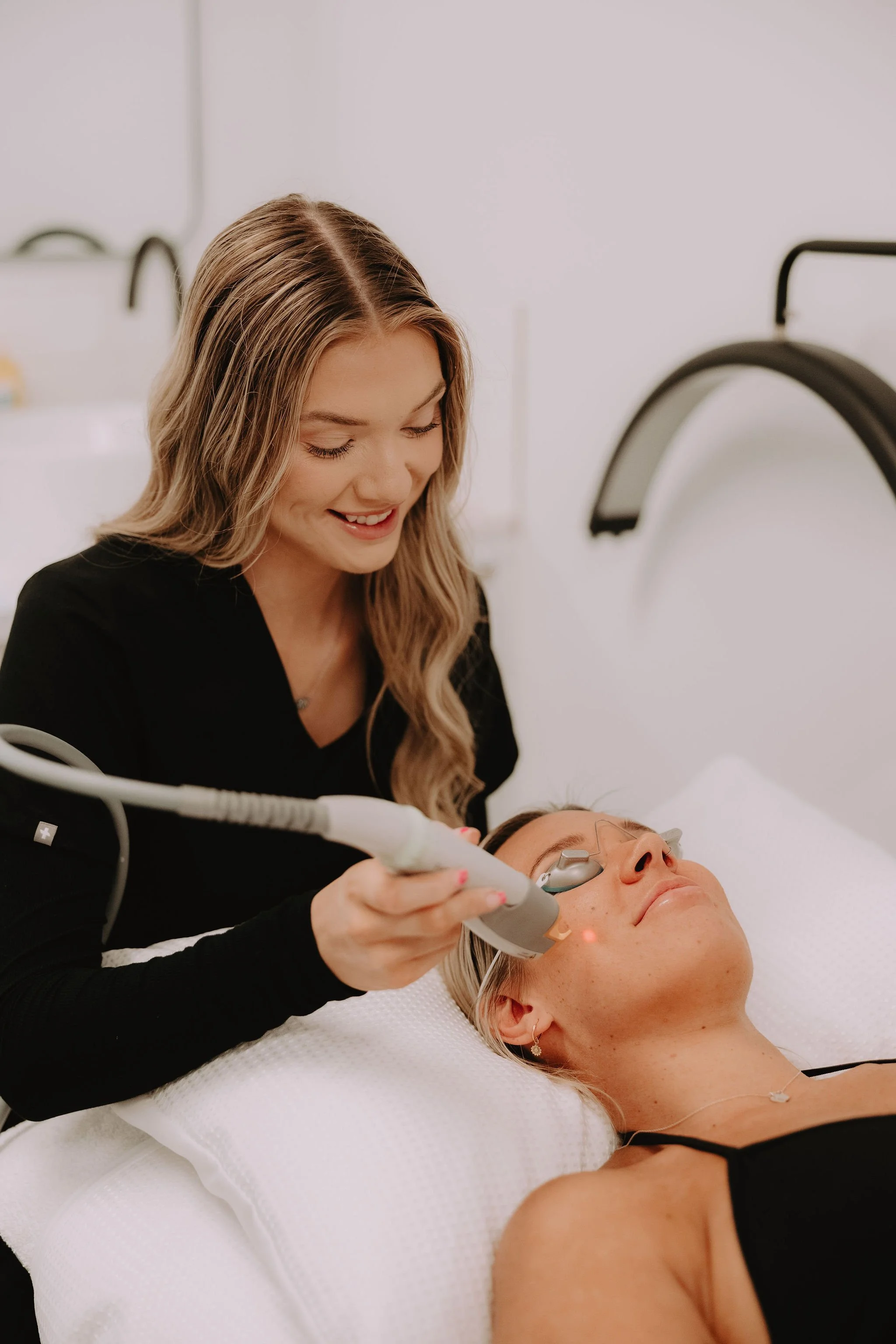 A woman with long blonde hair and a black top is performing a facial treatment with a handheld device on another woman lying down, who has blonde hair, wearing eye protection and a black top, in a medical setting.