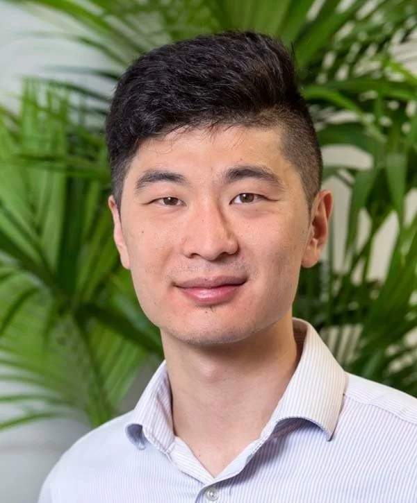A young Asian man with short black hair, wearing a light-colored dress shirt, standing in front of green tropical plants.