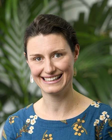 A woman smiling, wearing a blue floral dress and pearl earrings, with greenery in the background.