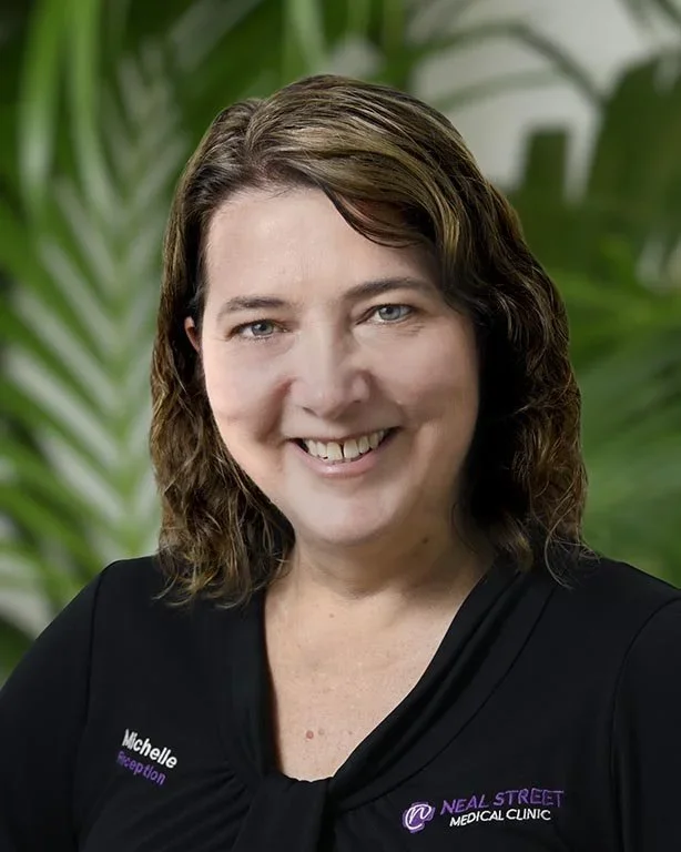 Smiling woman with shoulder-length brown hair, wearing a black medical scrubs top with "Michelle" embroidered on it and a background of green tropical plants.