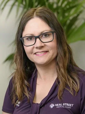 Portrait of a woman with brown hair and glasses, smiling, wearing a purple shirt with the Neal Street Medical Clinic logo, and standing in front of green plants.