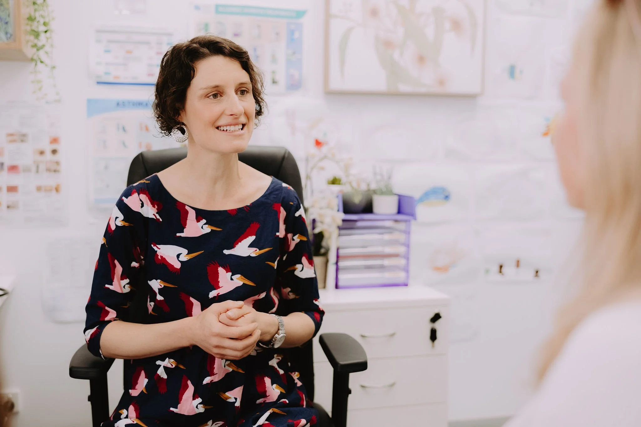A woman with short dark curly hair smiling and talking to another person in an office setting. She is wearing a navy dress with a pelican print and sitting on an office chair.