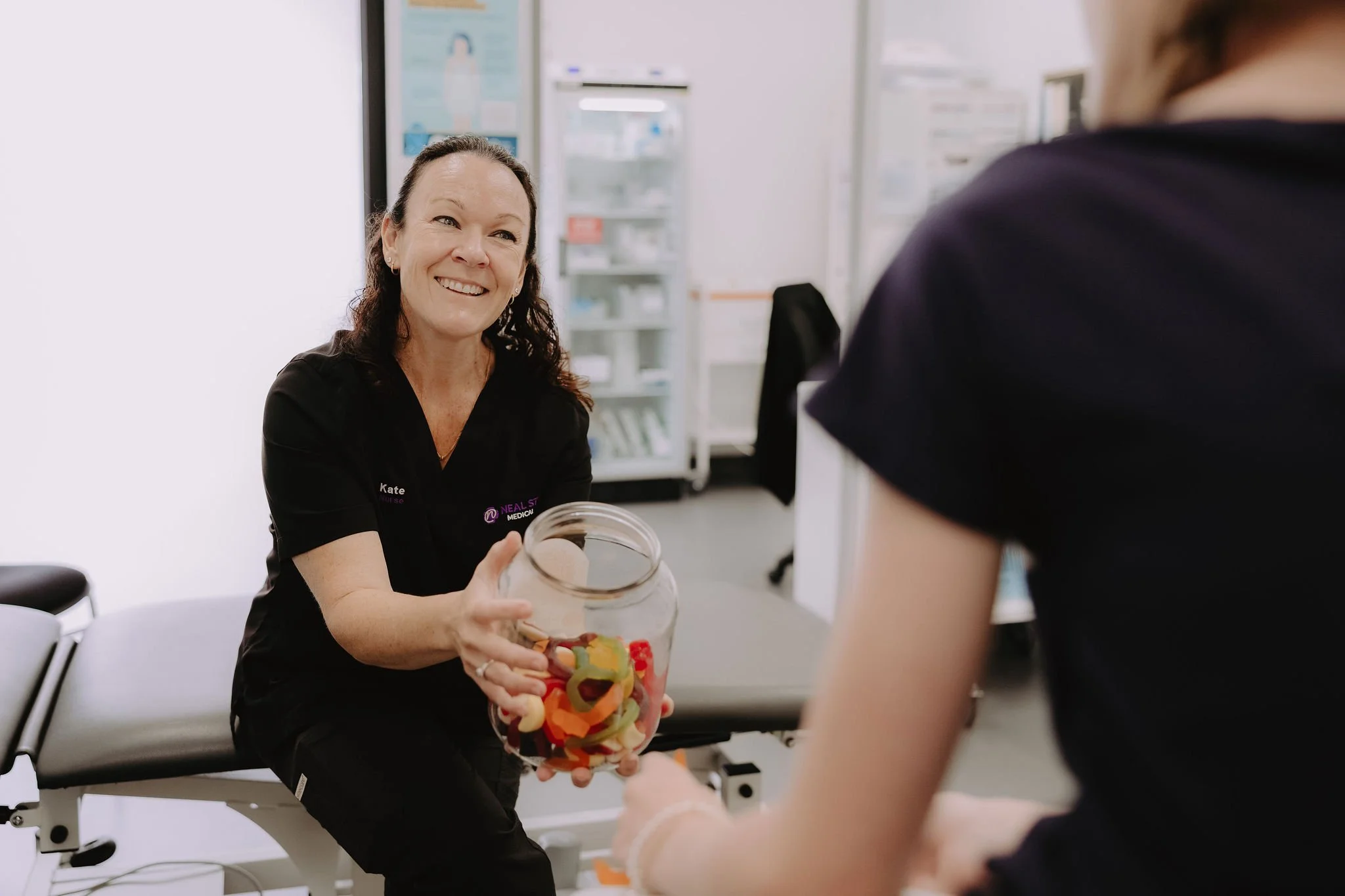 A healthcare worker smiling and handing a jar of colorful gummy bears to a patient in a medical office.