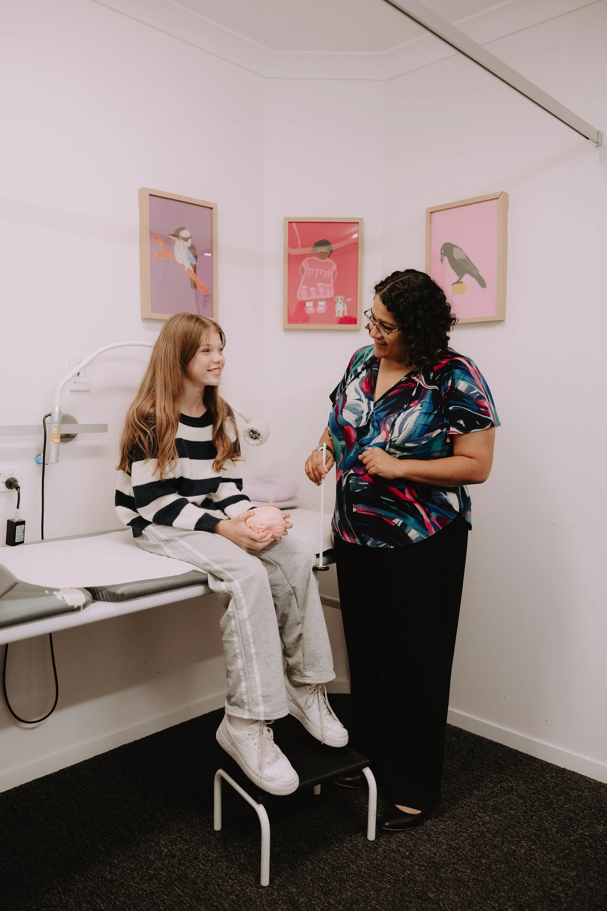 A young girl sitting on an examination table at the doctor’s office, holding a pink toy, smiling at the female healthcare professional standing beside her, in a room with colorful wall art.