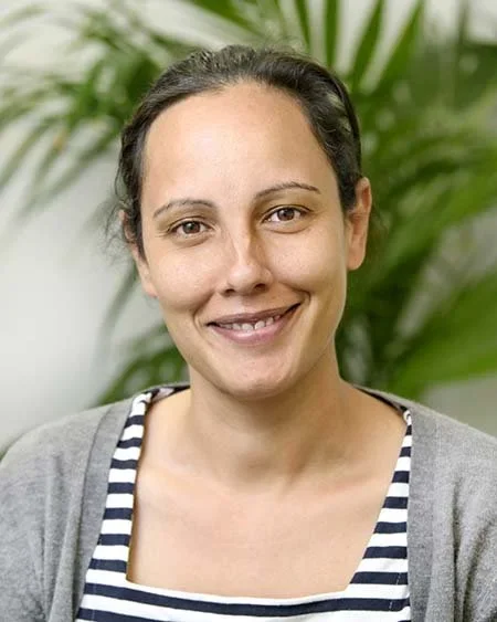A woman smiling with dark hair pulled back, wearing a striped shirt and gray sweater, outdoors with green plants in the background.