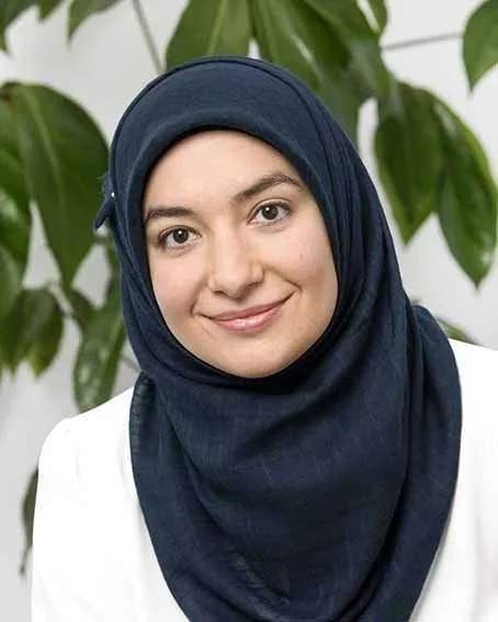 A woman wearing a navy blue hijab and white blazer, smiling in front of large green plant leaves.
