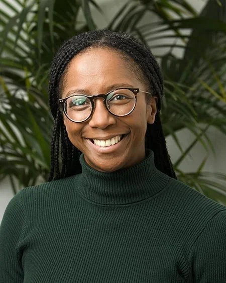 A woman with glasses and braided hair, smiling, standing in front of green leafy plants.