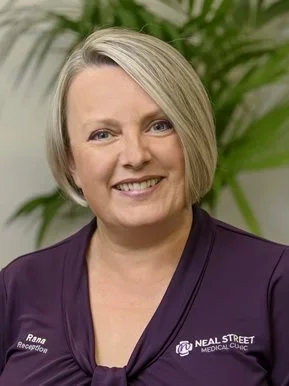 Portrait of a woman smiling in front of a plant, wearing a purple top with a badge that reads 'Rana Receptionist' and 'Neal Street Medical Clinic.'