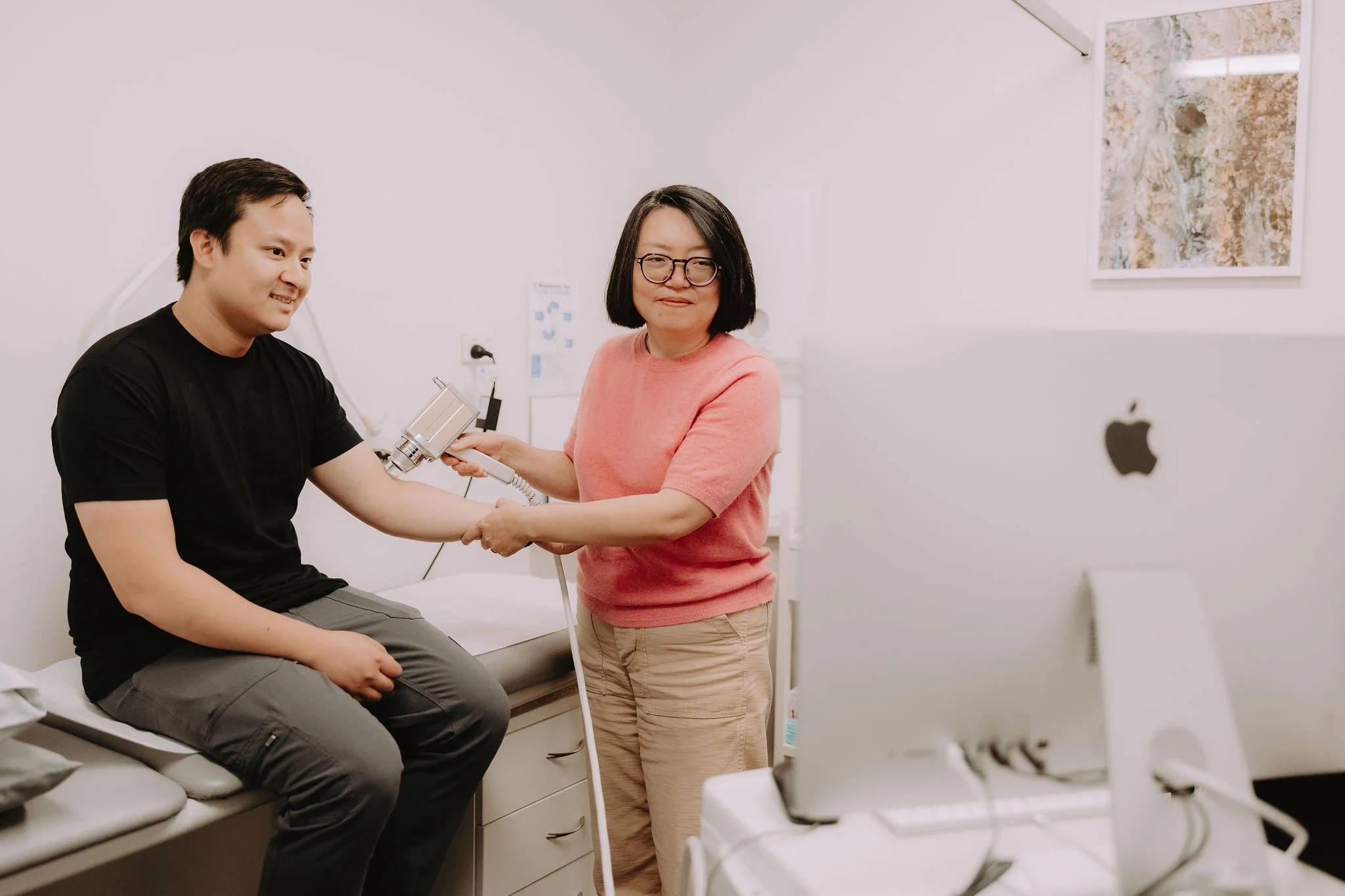 A woman holding a medical device is shaking hands with a young man sitting on an examination table in a medical room.