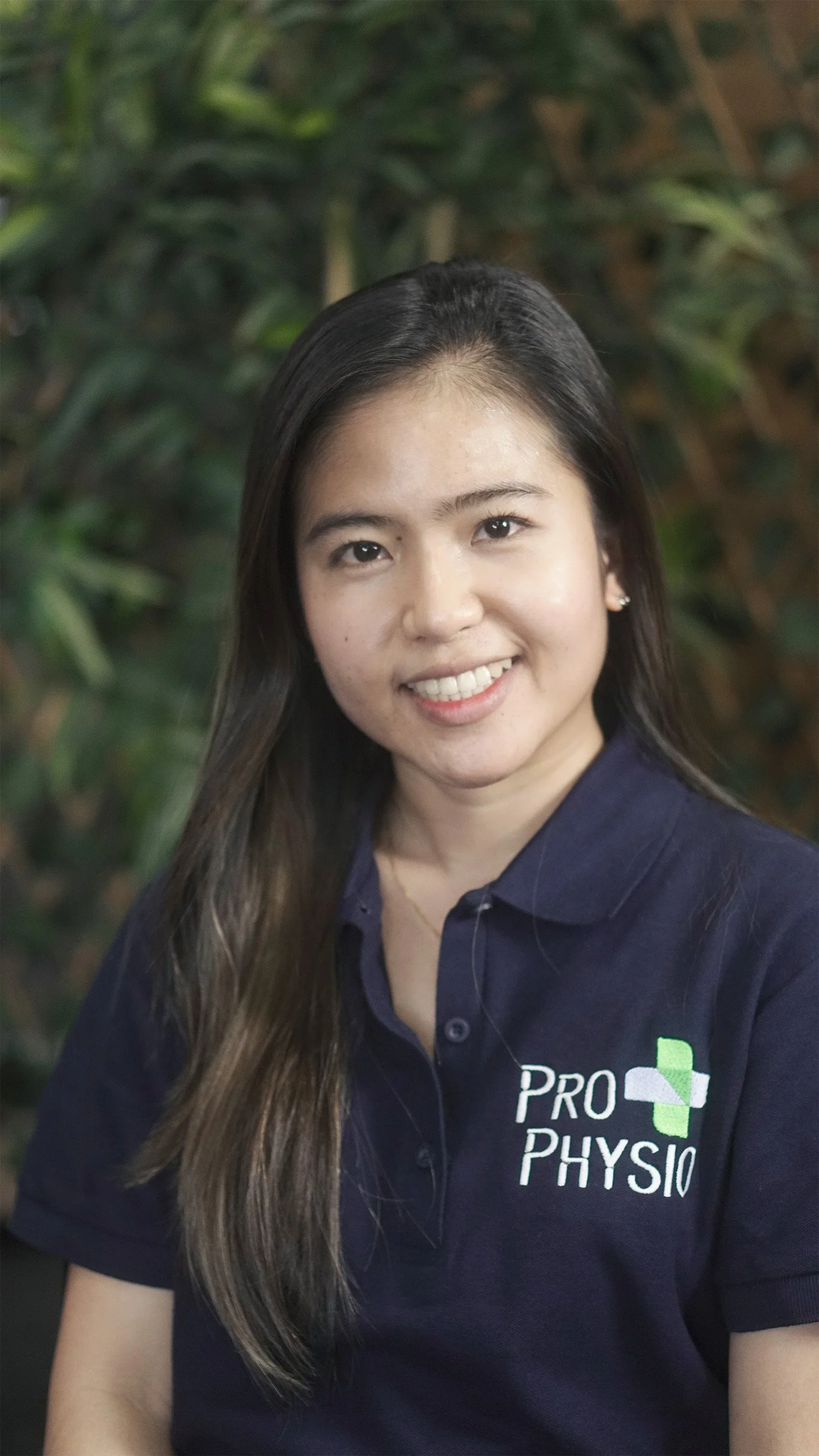 Portrait of a young woman with long dark hair, smiling, wearing a navy polo shirt with 'Pro Physio' logo, in front of green foliage