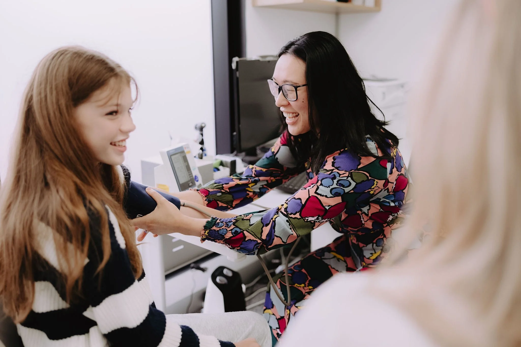 A healthcare professional, a woman with black hair and glasses wearing a colorful floral dress, using a stethoscope to listen to the heartbeat of a young girl with long red hair and a striped sweater, inside a medical office.