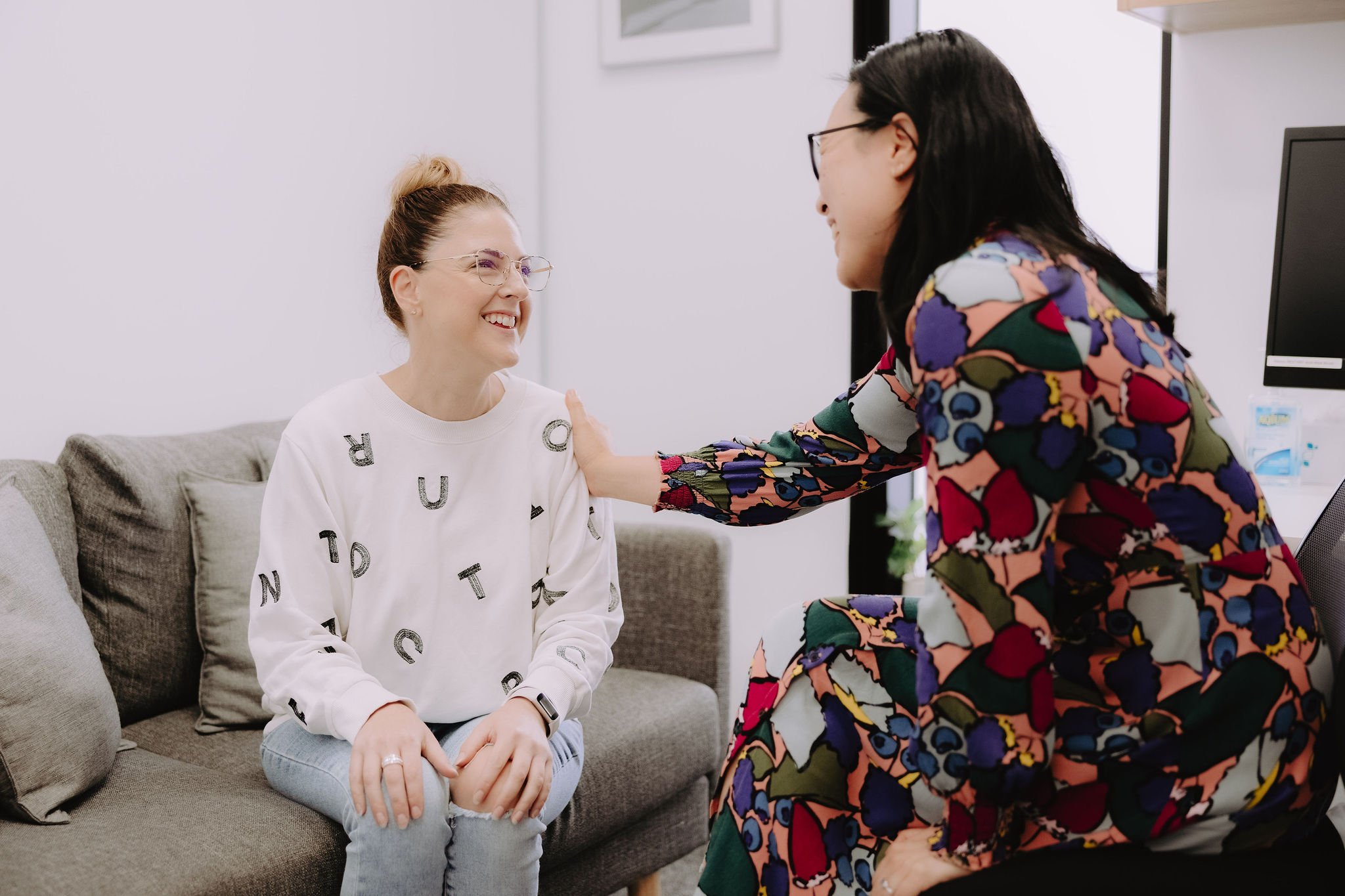 A woman with glasses and a patterned jacket is smiling and has her hand on the shoulder of a woman sitting on a sofa, who is also smiling and wearing glasses and a white sweater with black letters. They are in an office setting with a computer and picture frames on the wall.
